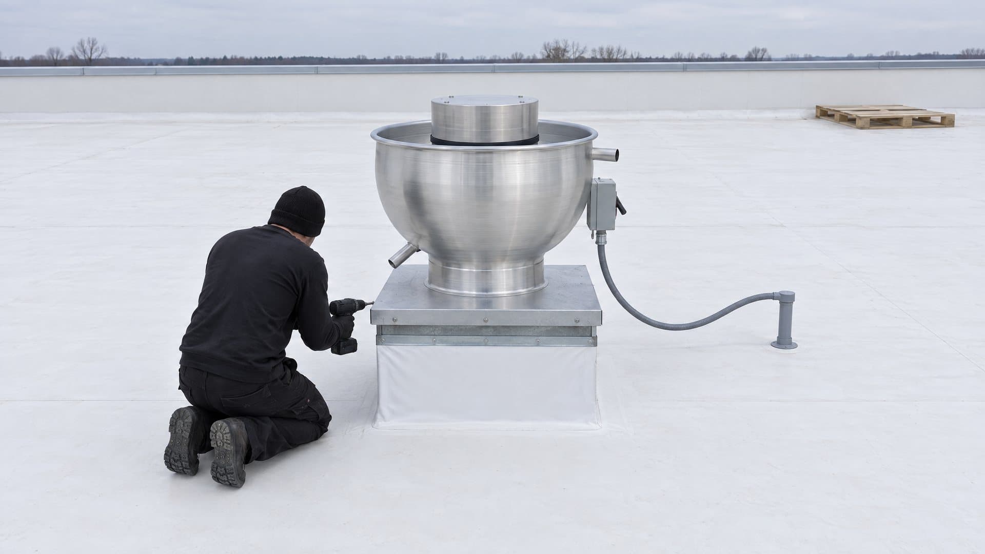 Technician kneeling on a white roof, screwing a new upblast exhaust fan's curb cap down onto its freshly flashed roof curb