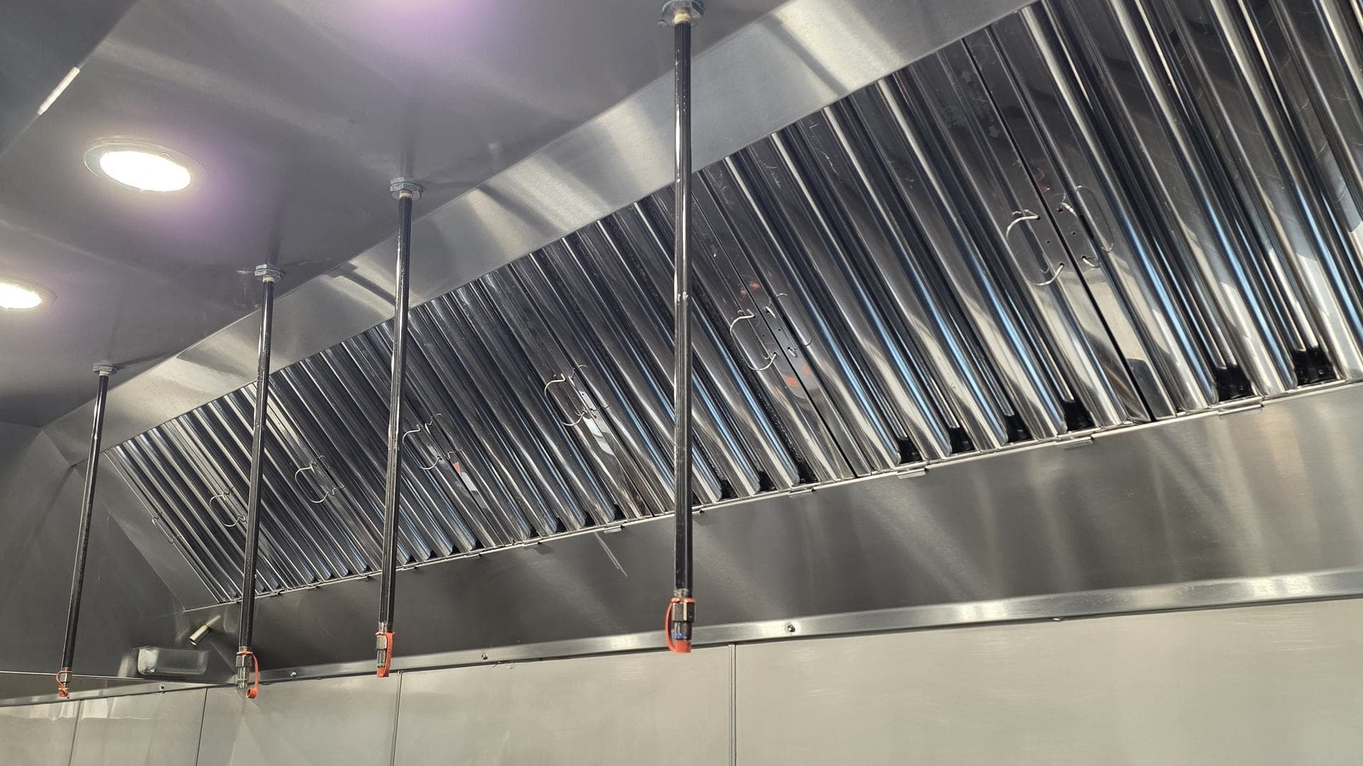 Wet-chemical fire suppression drops inside a cleaned kitchen hood, each nozzle under an orange blow-off cap, in front of bright baffle filters