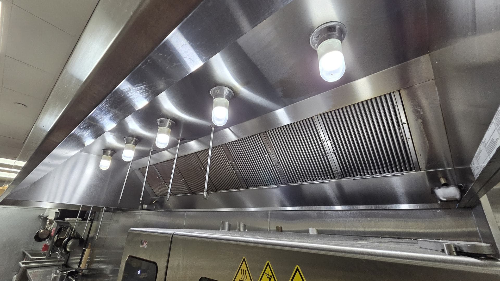Stainless exhaust hood over a restaurant cook line, with globe lights and baffle filters above the ovens