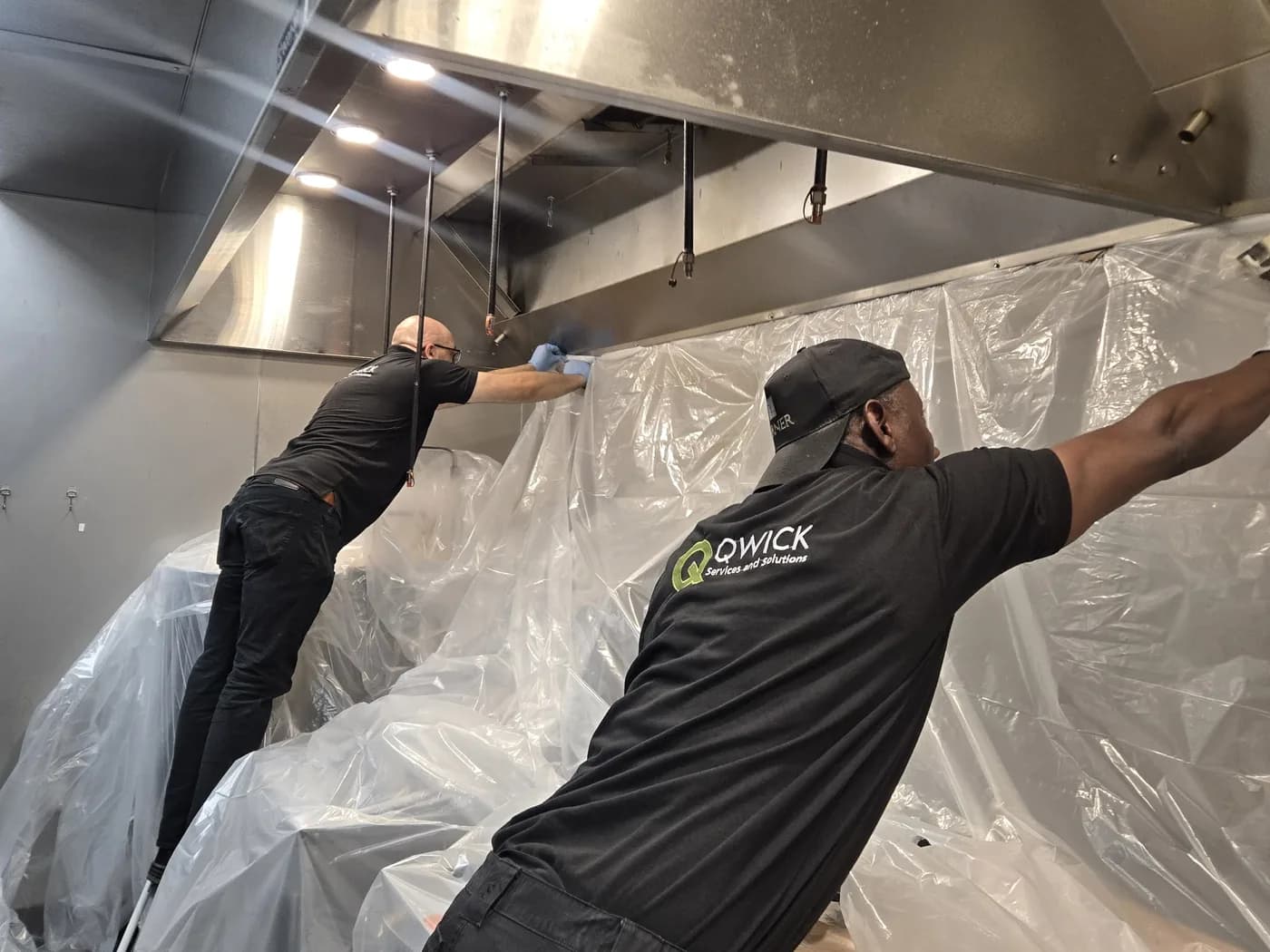 Two Qwick technicians taping clear plastic from a kitchen hood down over the cook line before a hood cleaning